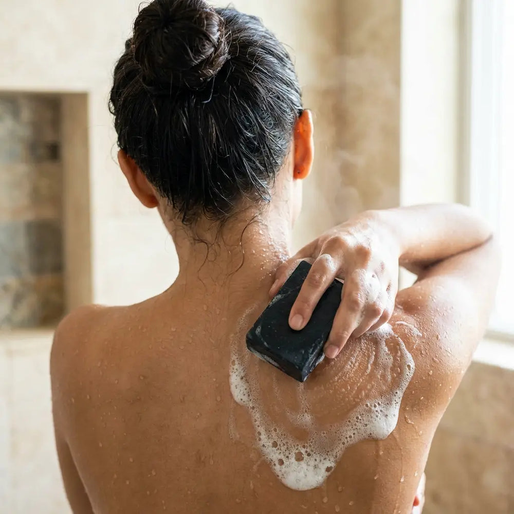 Woman in the shower reaching over her shoulder to work a dark soap bar across her upper back.