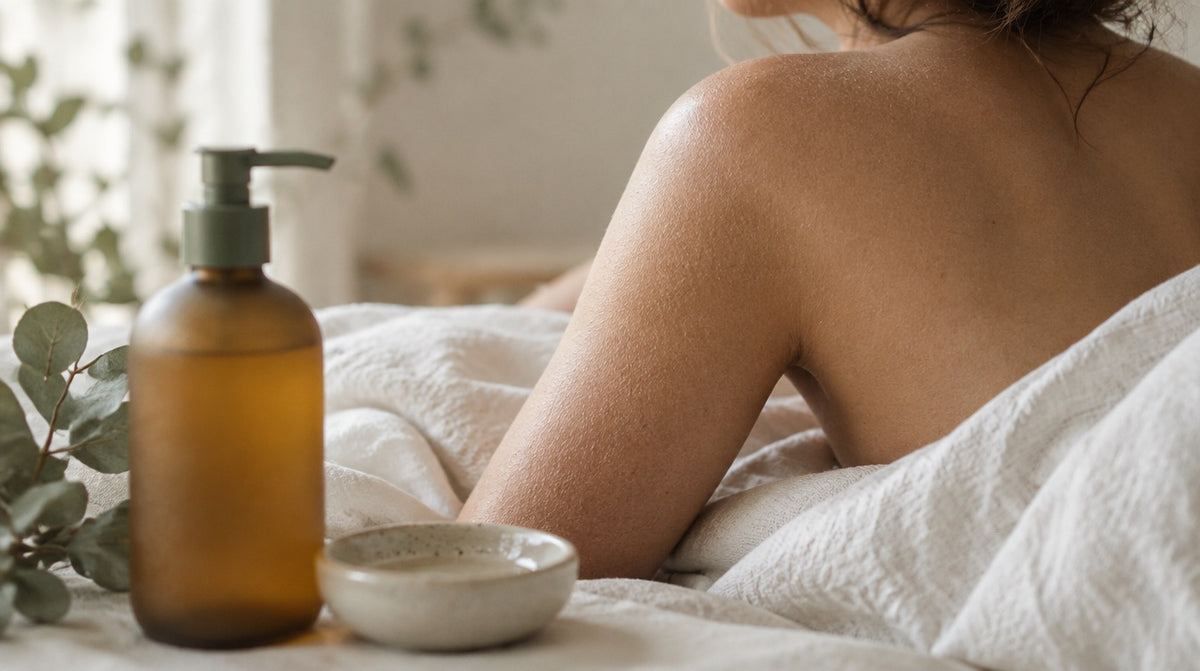 Woman's bare upper arm and shoulder seen from behind on white linen, with an amber glass body wash bottle and small ceramic dish in the foreground