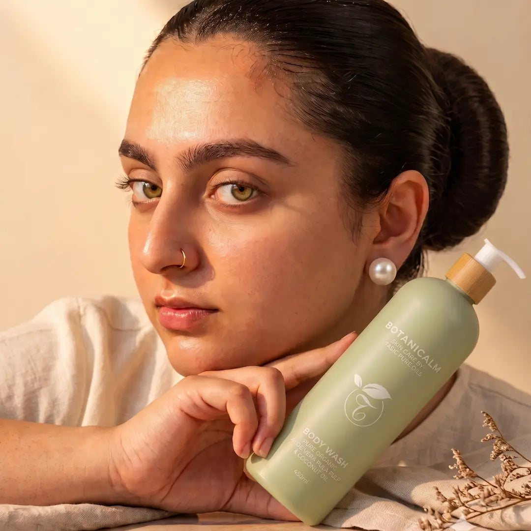 A woman with dark hair in a bun holds a bottle of Botanically Aligned Body Wash. She is wearing a white shirt and pearl earring, with a warm beige background and dried flowers in the foreground.
