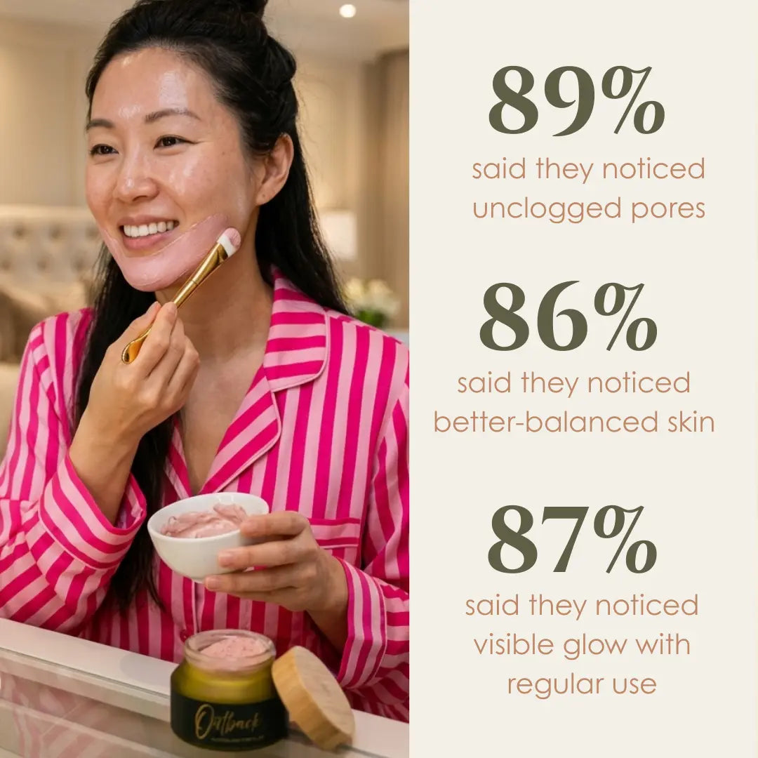 A smiling woman in pink striped pajamas applies a pink skincare cream from a small bowl with a gold-handled brush while standing in front of a mirror. A jar of the product sits on the counter below.