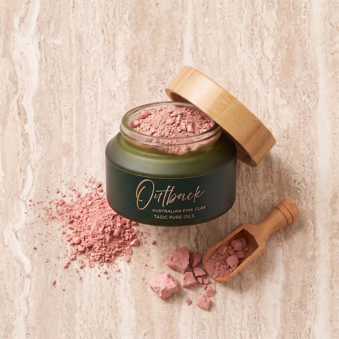 An open jar of the Tasic Pure Oils Outback Australian Pink Clay Mask with its bamboo lid propped beside it, on a light beige stone surface with scattered pink clay powder and a small wooden scoop.