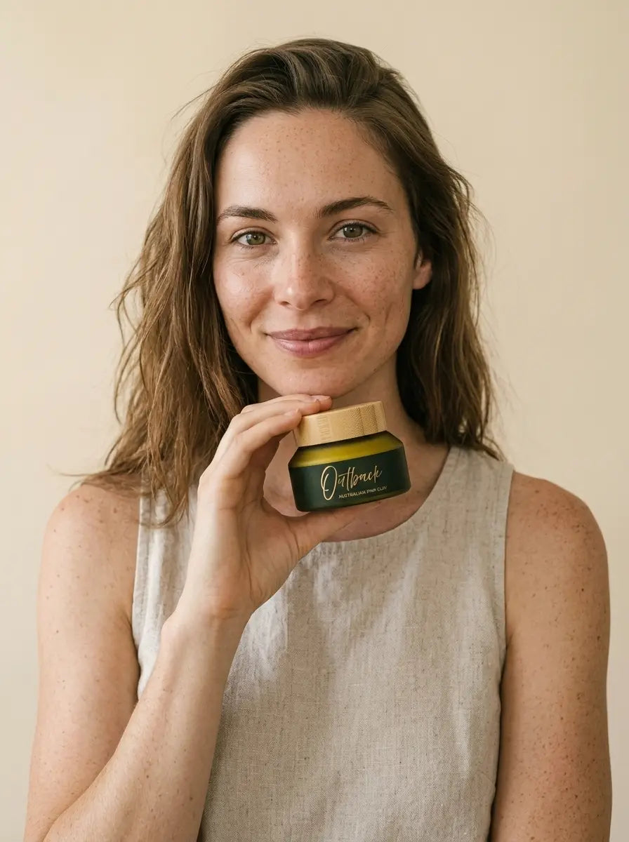 A woman with light brown hair holds a small green cosmetic jar with a gold lid in front of her face. The jar has the brand name 'Outbreak' printed on it.