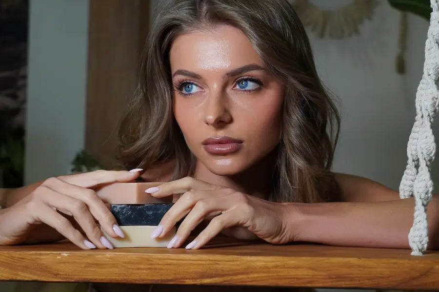 Woman with blue eyes and light brown hair holding a bar of soap on a wooden surface.