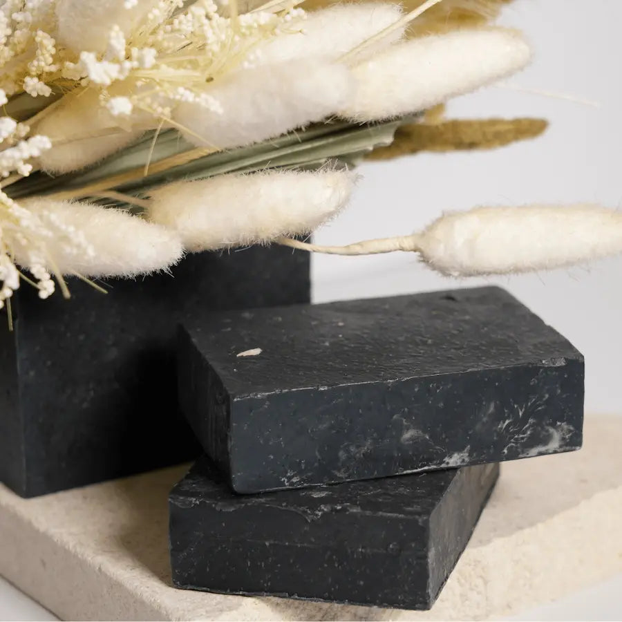 Three black soap bars on a light surface with decorative flowers in the background